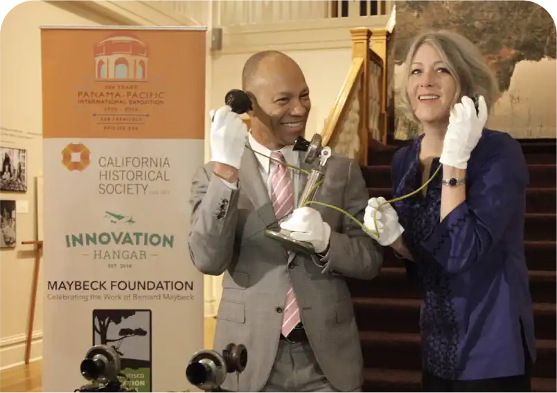 Ein Mann und eine Frau, beide mit weißen Handschuhen, lächeln, während sie die Hörer eines antiken Telefons in einer historischen Ausstellung halten.